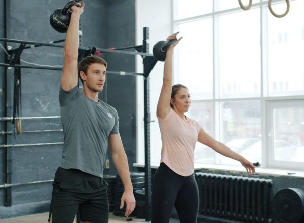 Young couple man and woman are training in gym weightlifting squatting with kettlebells together concentrated on bodybuilding exercises indoors.